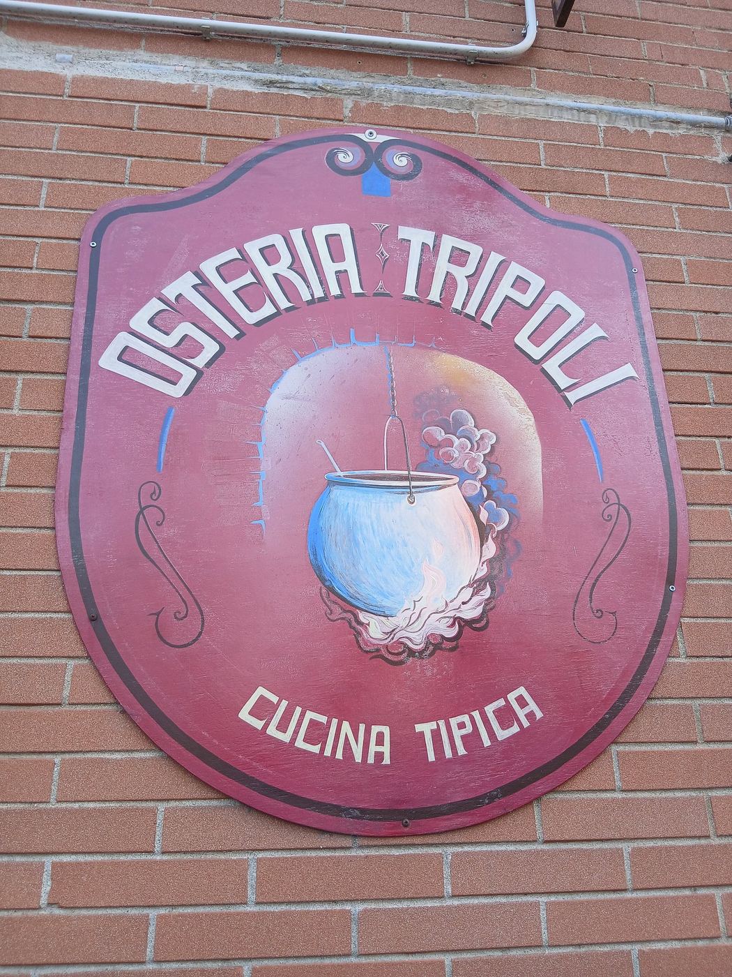 Insegna dipinta dell'Osteria Tripoli su muro di mattoni: scudo rosso con un paiolo e la scritta Cucina tipica